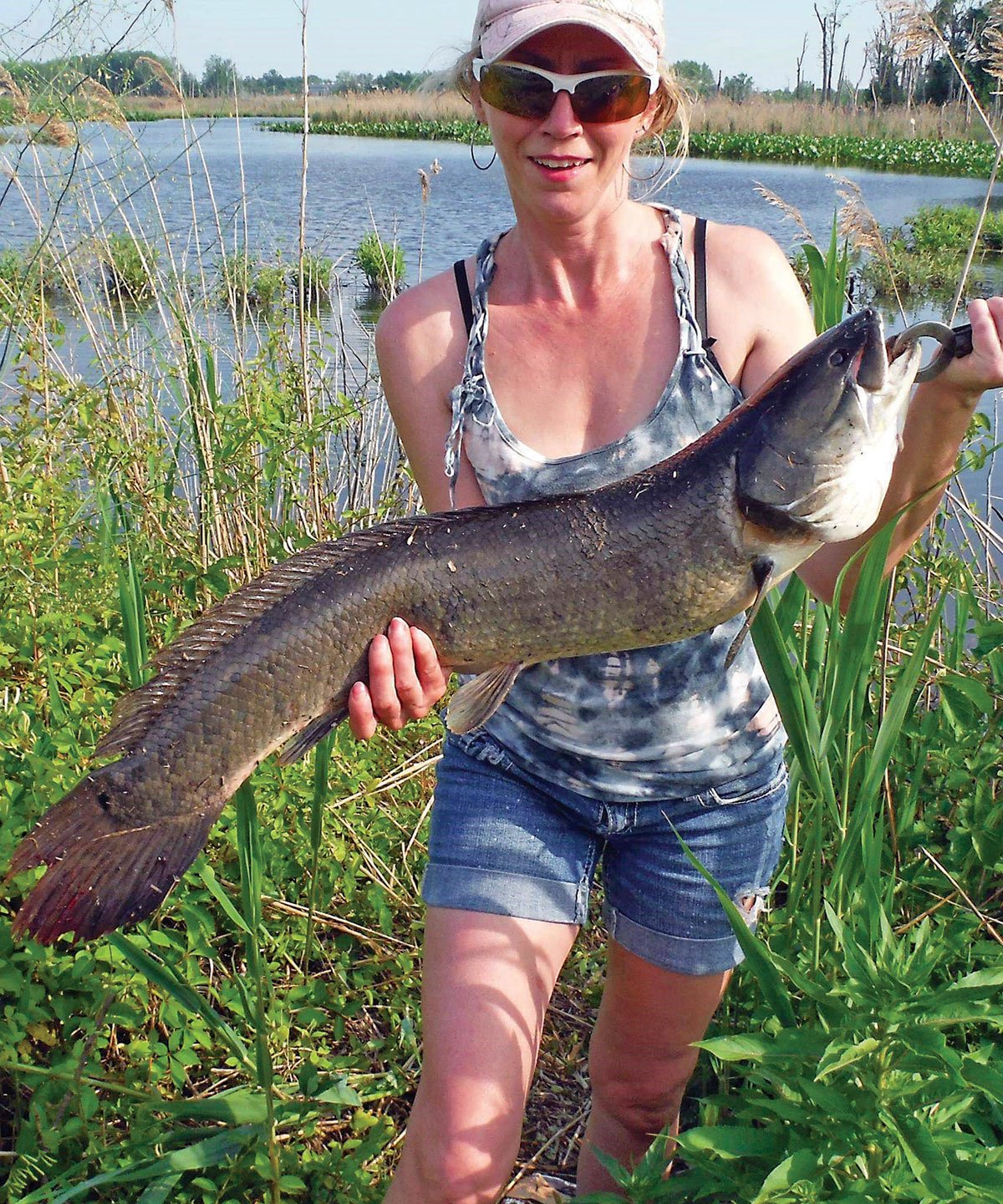 Woman in the grass near water holding a big bowfin fish