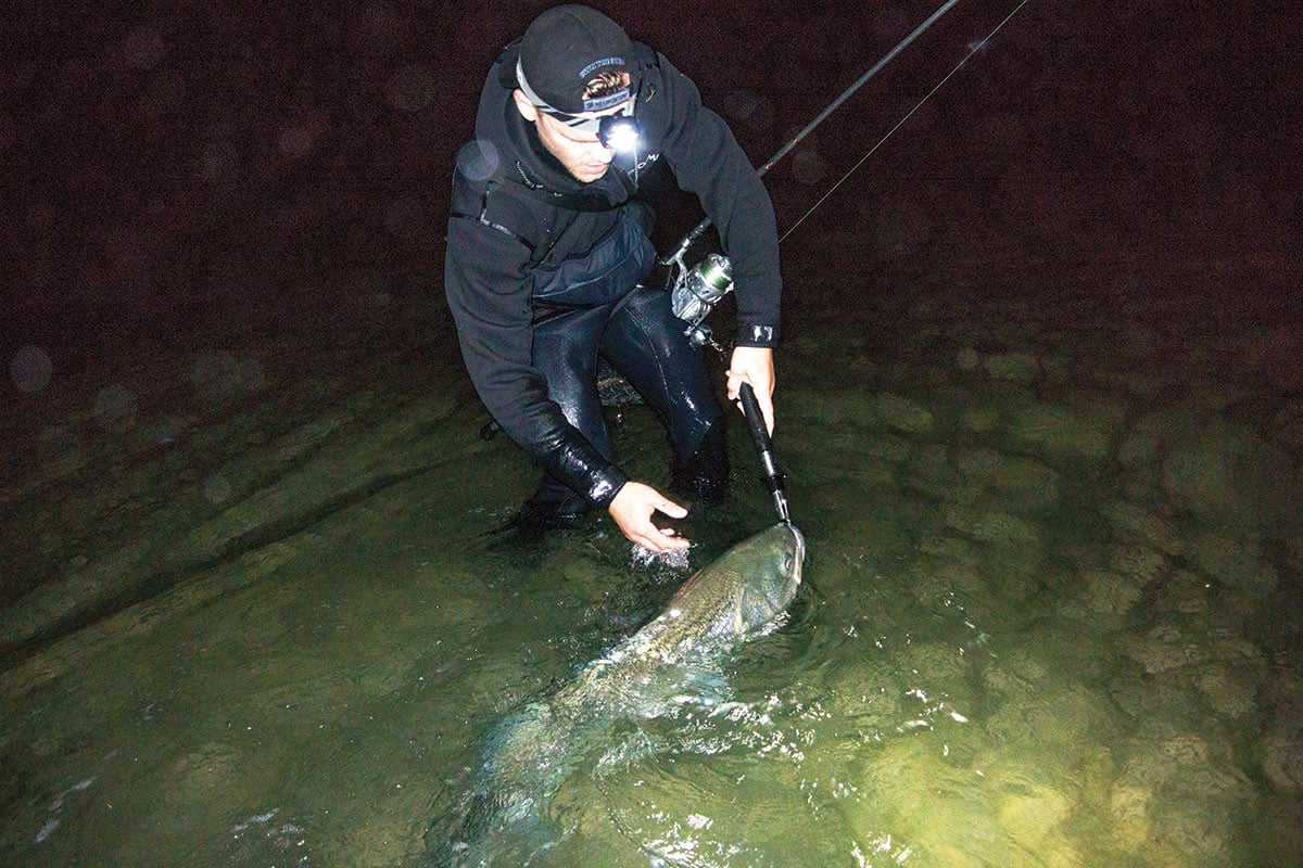 Man on knee deep water holding a fishing line with fish attached to the end